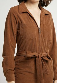 Jumpsuit en velours côtelé marron avec fermeture éclair à l'avant, deux poches poitrine, manches longues et ceinture à nouer à la taille. Tissu texturé en côtes.