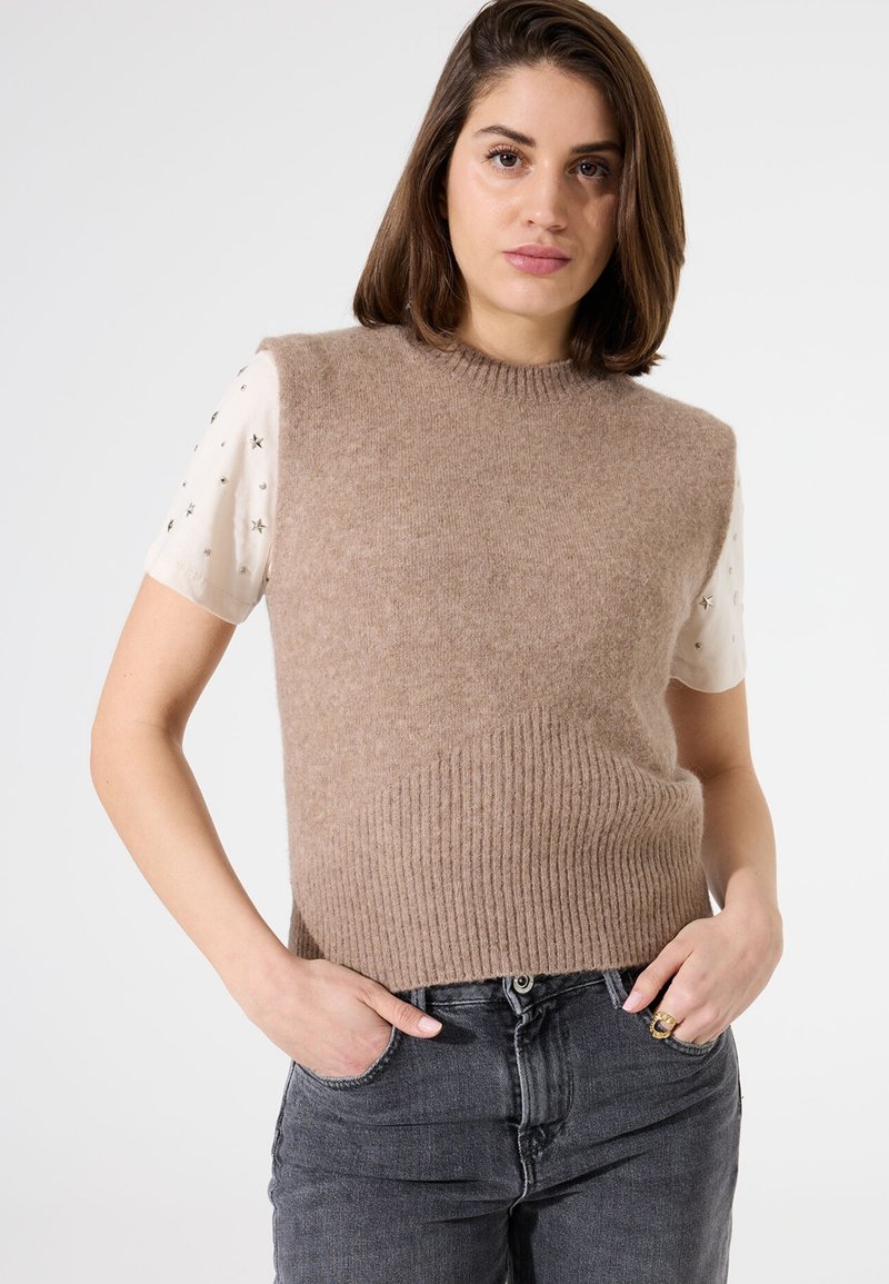 Brauner strickloser Pullover mit geripptem Saum und diagonalen Texturdetails, über einem kurzärmeligen weißen Oberteil mit Sternmustern getragen.