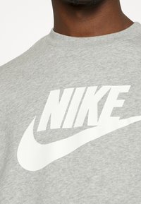 Grå sweatshirt med en stor vit Nike-logotyp och swoosh-design. Materialet verkar mjukt med en klassisk rund hals och korta ärmar.