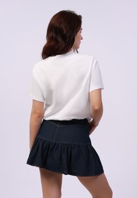 Camiseta de algodón blanca combinada con una falda de mezclilla oscura con un dobladillo acampanado y una cinturilla plana, vista desde atrás.