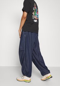 Tmavě modré baggy džíny s bočními kapsami, v kombinaci s černým tričkem s barevným potiskem na zádech a světlými teniskami.