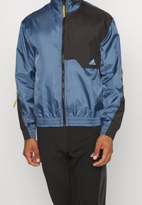 Blau- und schwarze Windbreaker-Jacke mit Reißverschluss vorne, elastischen Bündchen und Taillenbund. Mit goldenem Reißverschluss und Adidas-Logo auf der Brust.