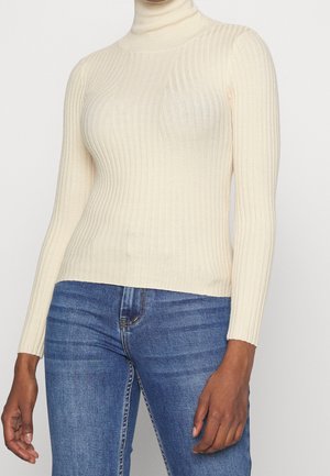 Personne portant un pull à col roulé côtelé crème et un jean en denim bleu, debout devant un fond neutre.