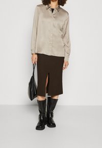 Seidene beigefarbene Bluse mit Knopfleiste, gerippter dunkelbrauner Midikleid mit Schlitz vorne, schwarze kniehohe Stiefel und eine schwarze Handtasche.