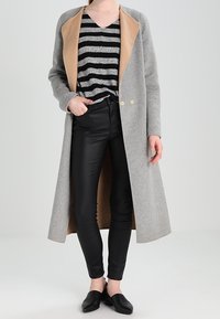 Grijze wollen jas met een tan kleurige kraag, dubbele knopen, gecombineerd met zwarte skinny jeans en een zwart-grijs gestreept top. Zwarte instap schoenen.