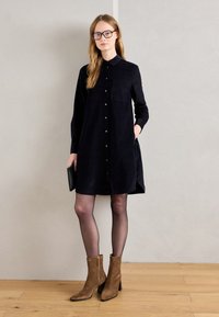 Donna con occhiali che indossa un vestito nero con bottoni, collant neri trasparenti e stivaletti alla caviglia in suede marroni, mentre tiene una pochette nera.