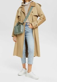 Manteau beige avec une capuche, doté de boutons croisés. Jean cropped bleu clair. Baskets blanches et un petit sac vert avec des poignées.