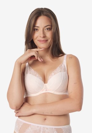 Kobieta o jasnobrązowych włosach, ubrana w bladoróżowy koronkowy biustonosz i pasujące do niego majtki, uśmiechająca się, z małymi tatuażami na ramionach i barku.