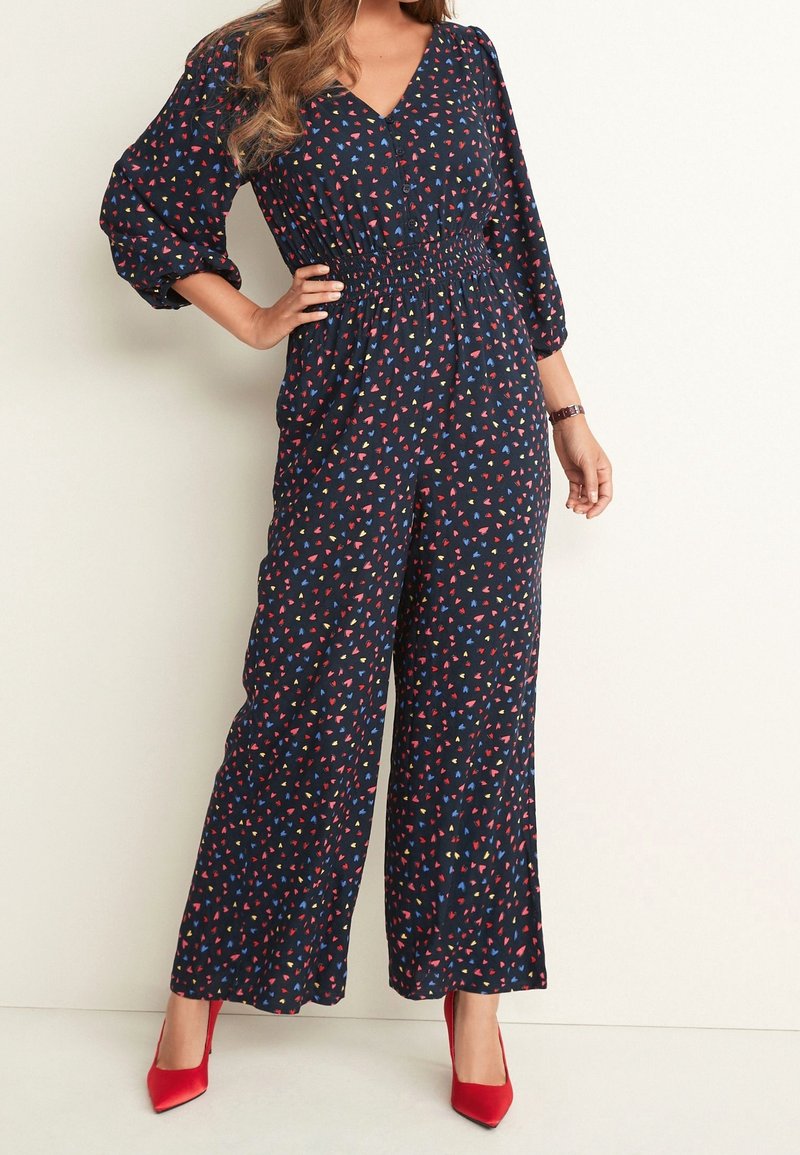 Femme portant une combinaison noire avec de petits motifs de cœurs colorés, des manches longues bouffantes et des talons hauts rouges pointus.
