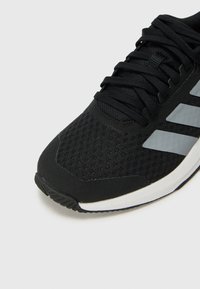Chaussure de sport noire avec une tige en mesh, rayures grises, et une semelle en caoutchouc. Présente des lacets plats et une finition texturée.