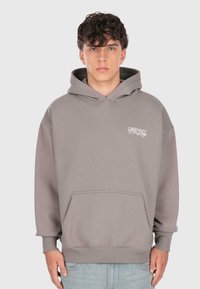 Mladý muž s kučeravými vlasmi, ktorý má na sebe hnedošedú mikinu s predným vreckom a malým bielym logom na hrudi, doplnenú o svetlomodré džínsy.