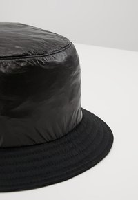 Pălărie neagră tip bucket, cu o textură lucioasă și crăpată. Are o margine lată și plată, precum și o coroană structurată. Confecționată dintr-un material neted.