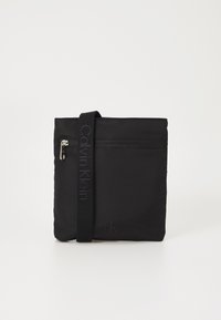 Sac à bandoulière en nylon noir avec une poche zippée, sangle en textile arborant la marque "Calvin Klein", et un logo en relief dans le coin inférieur.