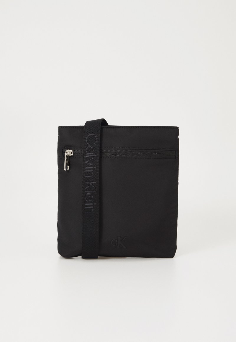 Sac à bandoulière en nylon noir avec une poche zippée, sangle en textile arborant la marque "Calvin Klein", et un logo en relief dans le coin inférieur.