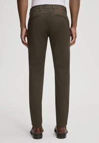 Pantaloni verde scuro con vestibilità slim, caratterizzati da una parte anteriore piatta, tasche posteriori e una texture liscia. Indossati con scarpe marroni.