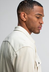 Beige overhemdjacket met een kraag, textuurstof en een borstzak met een donkere knoop. Zijaanzicht met stikdetails.