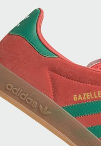 Tenisky Adidas Gazelle v červenej semišovej látke s zelenými akcentmi. Majú gumenú podrážku a vyrazené logo na boku. Klasický dizajn s nízkym strihom.