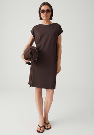 Donna che indossa un vestito marrone a maniche corte, occhiali da sole, infradito, che tiene una giacca marrone piegata abbinata contro uno sfondo chiaro e uniforme.
