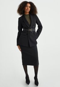 Črn eleganten blazer z dvoklopnim dizajnom, pasom za zožitev, ujemajočo svinčnico in vzorčastimi črnimi pantyhose.