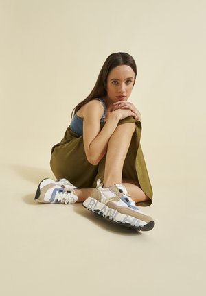 Jeune femme aux longs cheveux bruns assise par terre, serrant son genou, portant une jupe verte, un haut en denim et des baskets épaisses multicolores avec du texte sur les semelles.