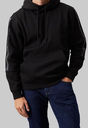 Mand iført sort hættetrøje med frontlomme og striber på ærmerne, kombineret med mørkeblå jeans, stående foran en ensfarvet baggrund.