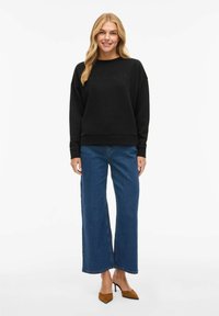 Smiling woman with long blonde hair wearing black sweatshirt with "MINIMALISM" text, blue wide-leg jeans, and brown pointed high heels.