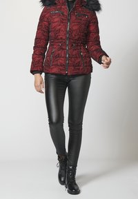 Osoba ubrana w czerwoną kurtkę w wzory z czarnym futrzanym kapturem, czarne skórzane spodnie i czarne sznurowane buty idzie do przodu.