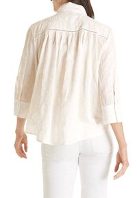 Helle cremefarbene Bluse mit Dreiviertelärmel, strukturiertem Stoff, zwei Knopfmanschetten und einer Rückenfalte. Verfügt über dekorative Stickerei am Ausschnitt.