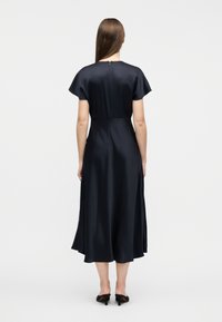 Γυναίκα με μακριά καστανά μαλλιά που φοράει μπλε-σκούρο μίντι φόρεμα με κοντά μανίκια, στέκεται με την πλάτη στραμμένη, φορώντας μαύρα mule με τακούνι.