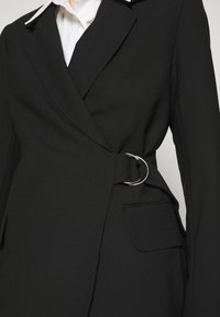 Blazer noir à design enveloppant, avec une boucle de ceinture circulaire argentée, une texture de tissu lisse et une poche latérale unique.