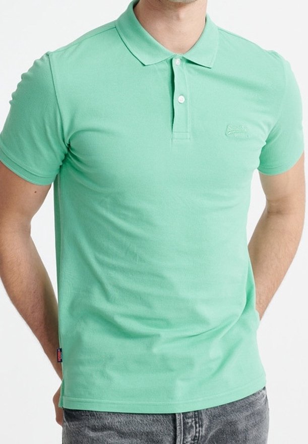 Homme portant un polo vert menthe à manches courtes avec deux boutons, associé à un jean gris, mains derrière le dos.