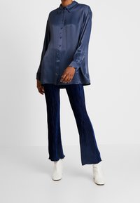 Marineblauw satijnen overhemd met knopen en lange mouwen, gecombineerd met plooibroek in marineblauw; model draagt witte enkellaarsjes met hak.