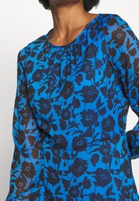 Kvinna iklädd en blå långärmad blus med svart blomsterprint och rynkad halsringning, sedd från axlarna till midjan.