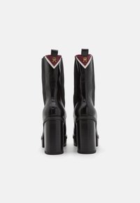 Zwarte leren enkellaarsjes met een hoge blokhak, voorzien van bordeauxrode accenten en een driehoekig wit paneel bij de hak met een gouden logo.