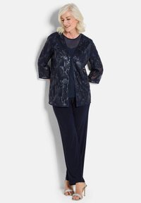 Marineblaue Jacke mit Dreiviertelärmeln und Paillettenstruktur über einem einfarbigen marineblauen Oberteil, kombiniert mit fließenden marineblauen Hosen und silbernen Absatzsandalen.