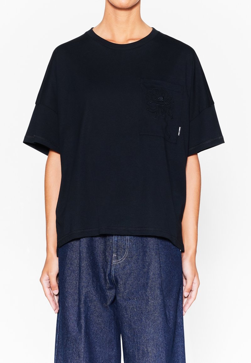 Camiseta de algodón negra con un bolsillo en el pecho que presenta un diseño floral bordado, de manga corta y corte relajado. Se lleva con pantalones de mezclilla.