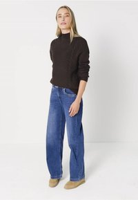 Ruskea palmikkoneule, jossa on korkea kaula-aukko, yhdistettynä sinisiin leveälahkeisiin farkkuihin ja beigekseen nilkkakorkokenkiin. Yksinkertainen valkoinen tausta kontrastina.