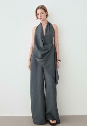 Kvinde står op ad hvid væg iført ærmeløs mørkegrå draperet haltertop og vidde bukser med armbånd om håndleddet.