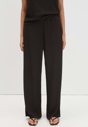 Person wearing loose black textured pants and matching sleeveless top, standing with hands relaxed by sides on plain background.