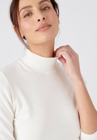 Top colore crema con collo alto e maniche lunghe, realizzato in materiale morbido e flessibile. Presenta una silhouette aderente senza motivi o dettagli.