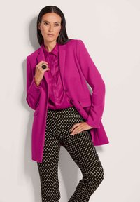 Fuchsia blazer met satijnen afwerking, zwart-witte gemusterde broek en een ring in de vorm van een roos. Gladde textuur, op maat gemaakt ontwerp.