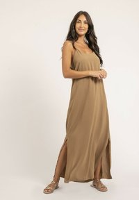 Robe maxi marron avec une texture lisse, bretelles spaghetti et fentes latérales hautes. Associée à des sandales argentées. Design simple et fluide.