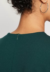 Temno zelena bluzica z gladko teksturo, okroglo ovratnikom in diskretnim zadrgo na zadnjem delu vratu.