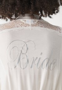 Grembiule bianco con dettagli in pizzo sulle spalle, caratterizzato da una stampa metallica argento "Sposa" sul retro in un carattere corsivo.