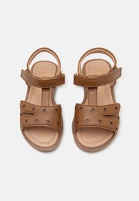 Braune synthetische Sandalen mit zwei verstellbaren Riemen und sternförmigen Ausschnitten. Weiches Fußbett, flache Sohle und ein Reißverschluss an der Ferse.