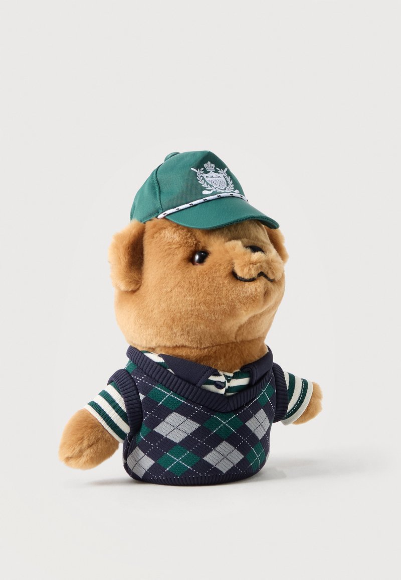 Plüschteddy mit grüner Mütze, gestreiftem Hemd und blau-grün kariertem Pullover, leicht nach rechts blickend vor einfachem Hintergrund.