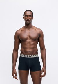 Czarne boxerki z szarym elastycznym pasem, na którym umieszczony jest napis "HILFIGER". Model przyjmuje neutralną pozę na tle czystego białego koloru.