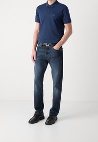 Námořnická modrá polo tričko s límečkem a malým logem, spárované s tmavě modrými džíny a černými botami. Outfit doplňuje černý opasek.