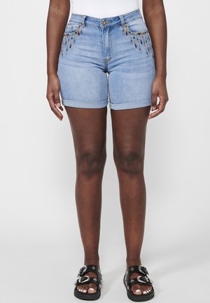 Femme portant un short en jean bleu clair avec des détails de perles colorées aux poches, un haut blanc et des sandales noires avec de grandes boucles argentées.