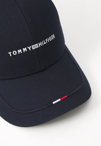 Marineblå kasket med en buet skygge. Har hvid "TOMMY HILFIGER" logo brodering og en rød, hvid og blå accent langs skyggen.